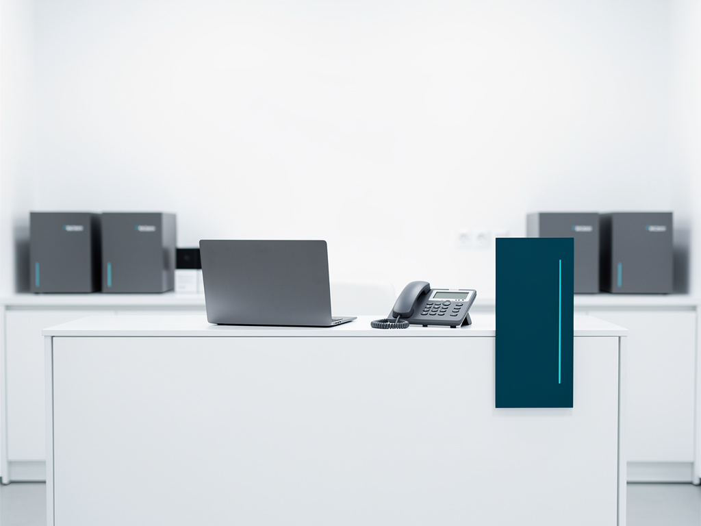 A minimal white front desk with a laptop, a phone and a small petrol-blue accent panel, nobody in focus.